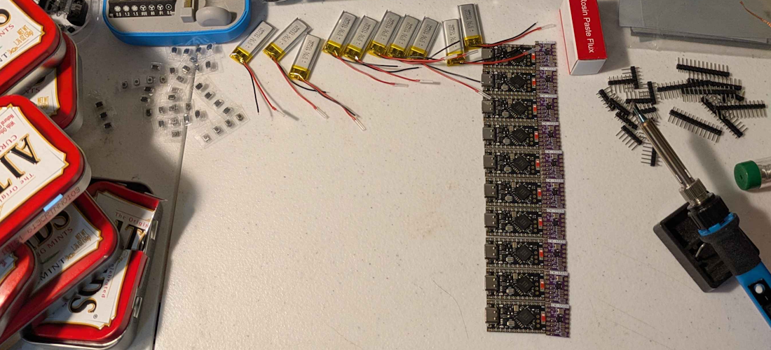 A folding table setup as a workbench for soldering and assembling components, with Altoids tins to the left, batteries above, and chips to the right