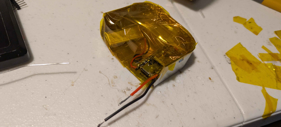 Half an Altoids tin, surrounded in yellow Kapton tape, with a black and red wire poking out from the side